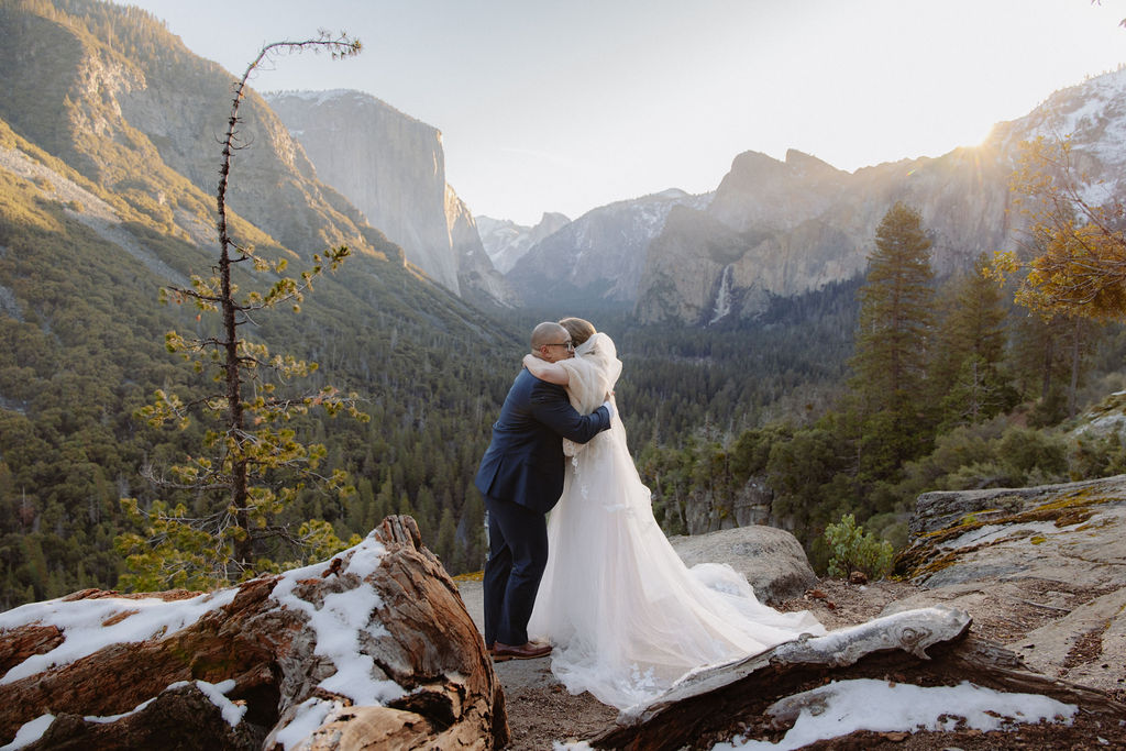 A bride and groom on a rocky ledge with mountains and forest in the background, under daylight for their sunrise yosemite elopement