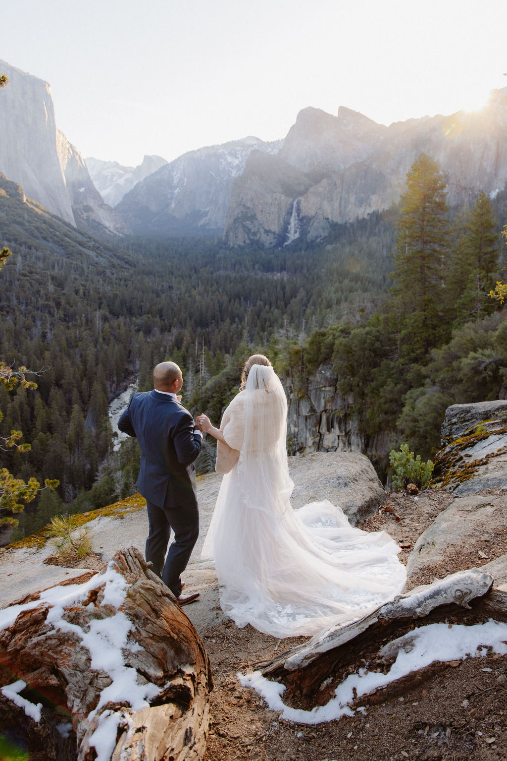 A bride and groom on a rocky ledge with mountains and forest in the background, under daylight for their sunrise yosemite elopement
