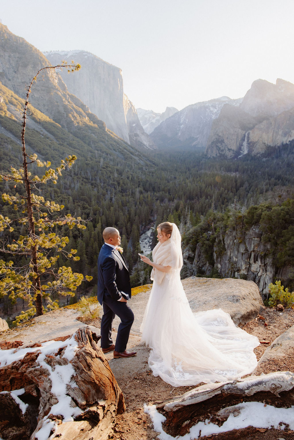 A bride and groom on a rocky ledge with mountains and forest in the background, under daylight for their sunrise yosemite elopement