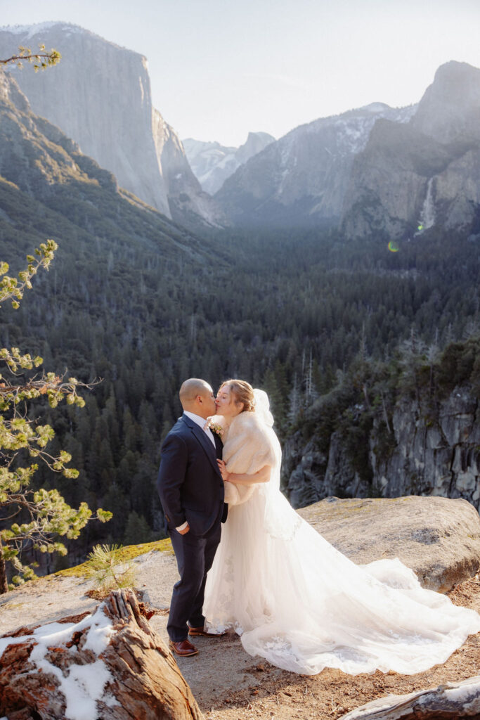 A bride and groom kiss on a rocky ledge with mountains and forest in the background, under daylight for their sunrise yosemite elopement
