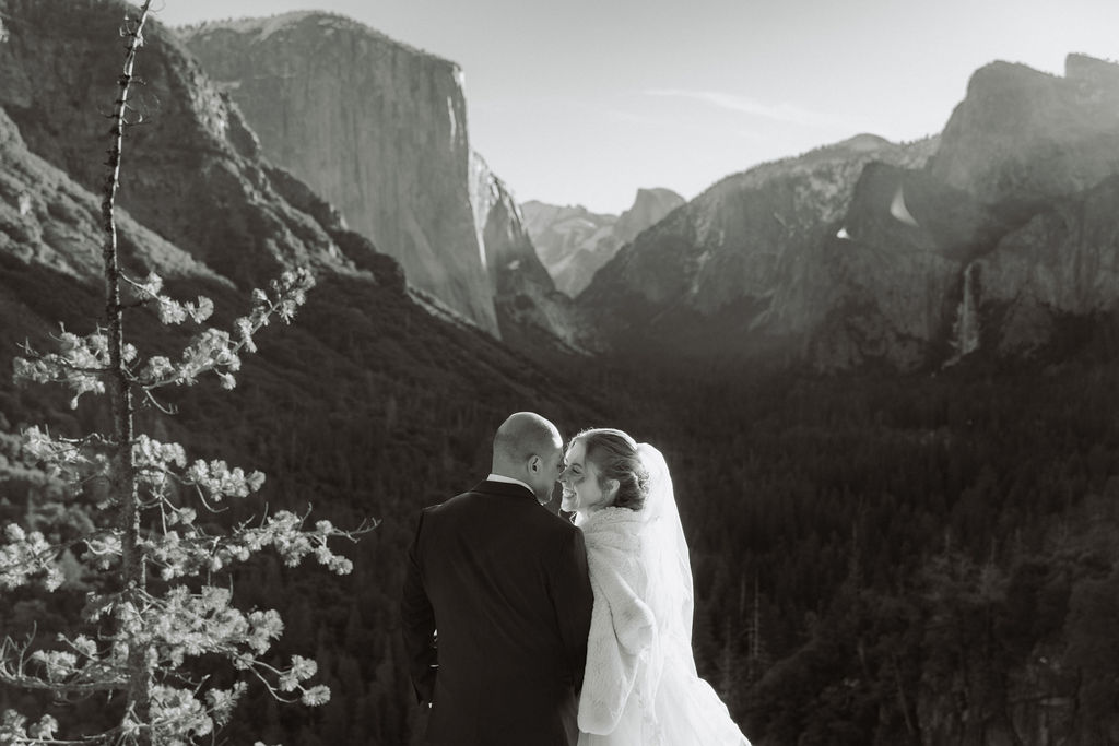 A bride and groom on a rocky ledge with mountains and forest in the background, under daylight for their sunrise yosemite elopement