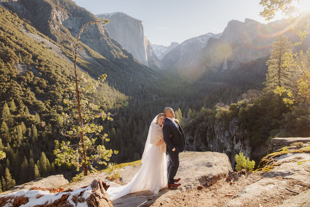 A bride and groom on a rocky ledge with mountains and forest in the background, under daylight for their sunrise yosemite elopement