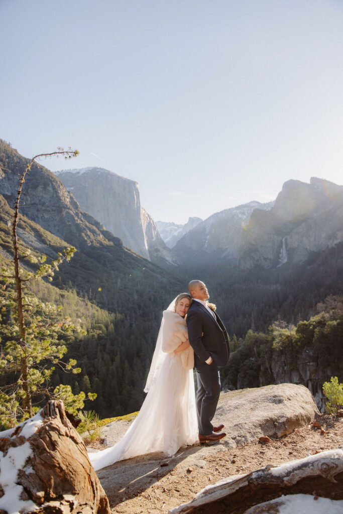A bride and groom kiss on a rocky ledge with mountains and forest in the background, under daylight for their sunrise yosemite elopement