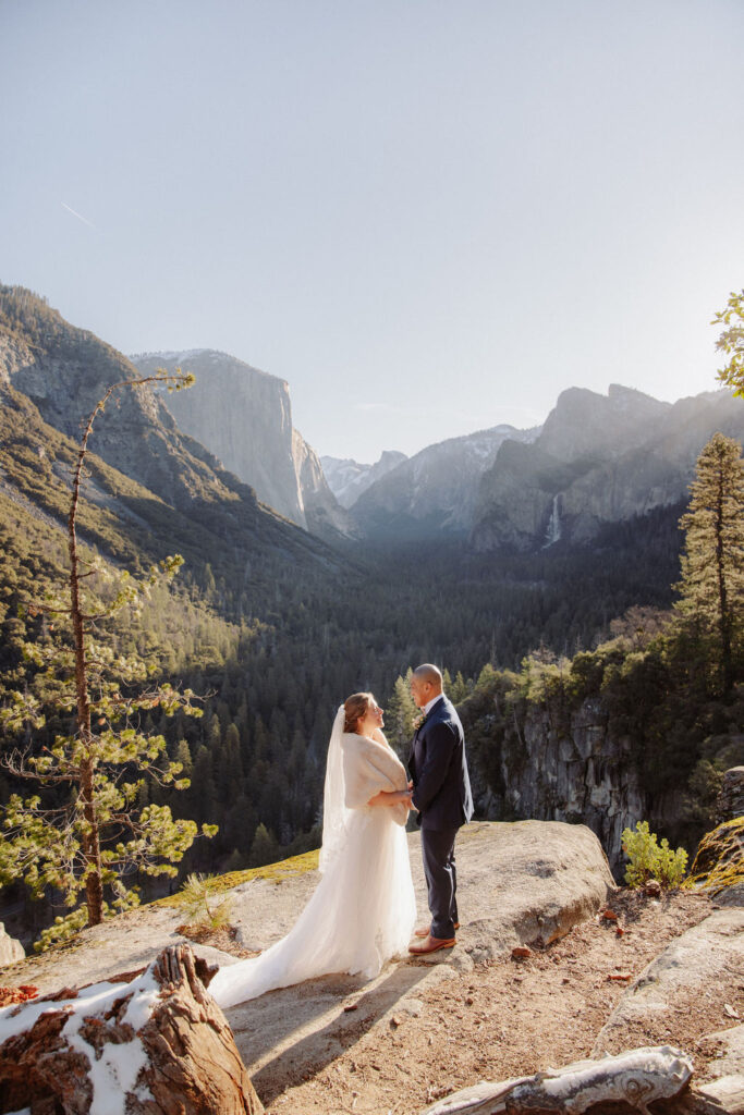 A bride and groom kiss on a rocky ledge with mountains and forest in the background, under daylight for their sunrise yosemite elopement