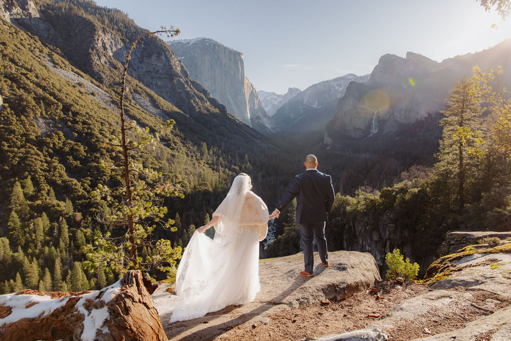 A bride and groom kiss on a rocky ledge with mountains and forest in the background, under daylight for their sunrise yosemite elopement