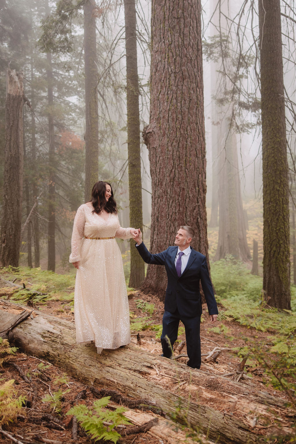 A woman in a long light dress stands on a fallen log in a forest while a man in a suit helps her balance. Tall trees and fog surround them.