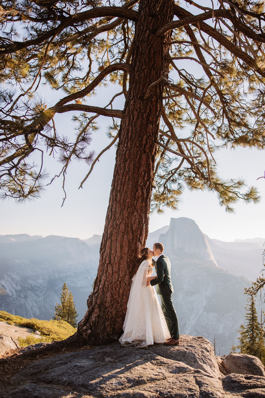 A bride and groom stand together on a rocky ledge with Half Dome mountain in the background under a clear sky.