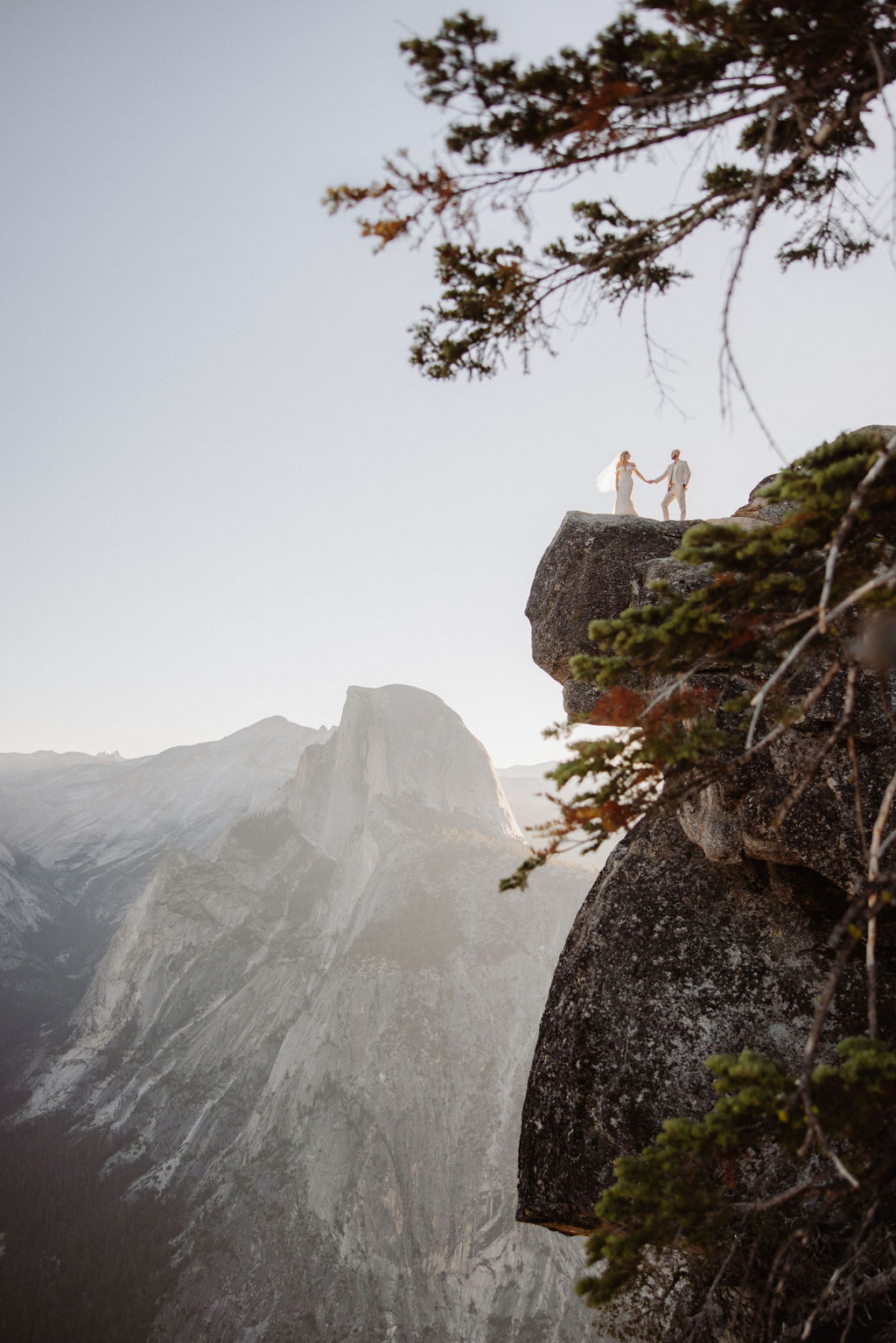 A person stands on the edge of a rocky cliff with mountains and trees in the background under a clear sky.