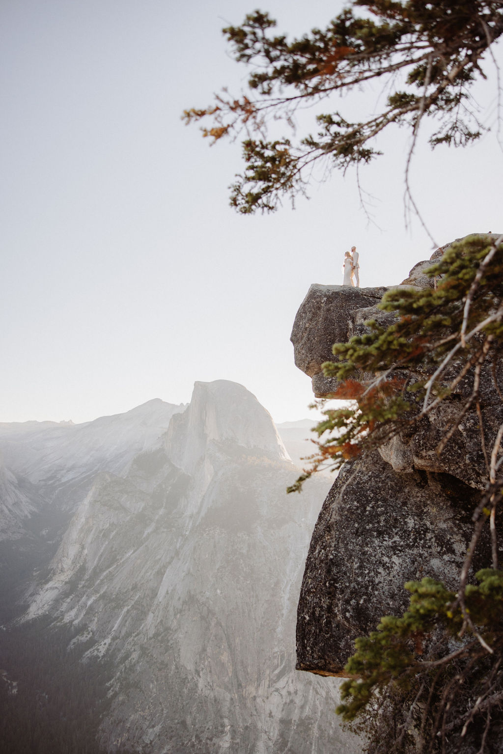 A person stands on the edge of a rocky cliff with mountains and trees in the background under a clear sky.