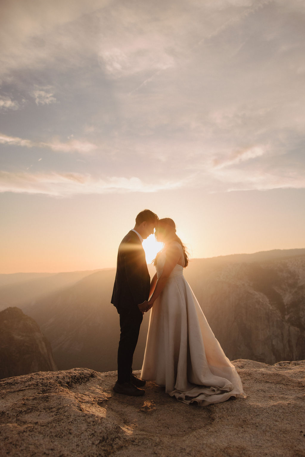 A bride and groom stand on rocky terrain, holding hands and facing a scenic mountain view at sunset. The Best Time for a Yosemite Elopement