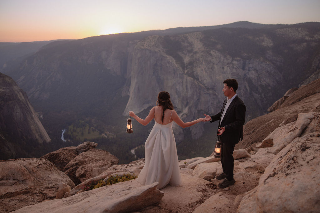 A bride and groom stand on rocky terrain, holding hands and facing a scenic mountain view at sunset. The Best Time for a Yosemite Elopement