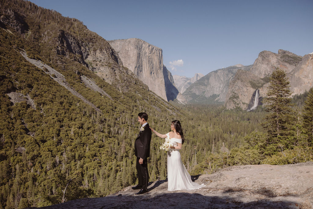 A bride in a white dress stands behind a groom in a black suit on a rocky overlook with mountains and trees in the background for their adventure elopement