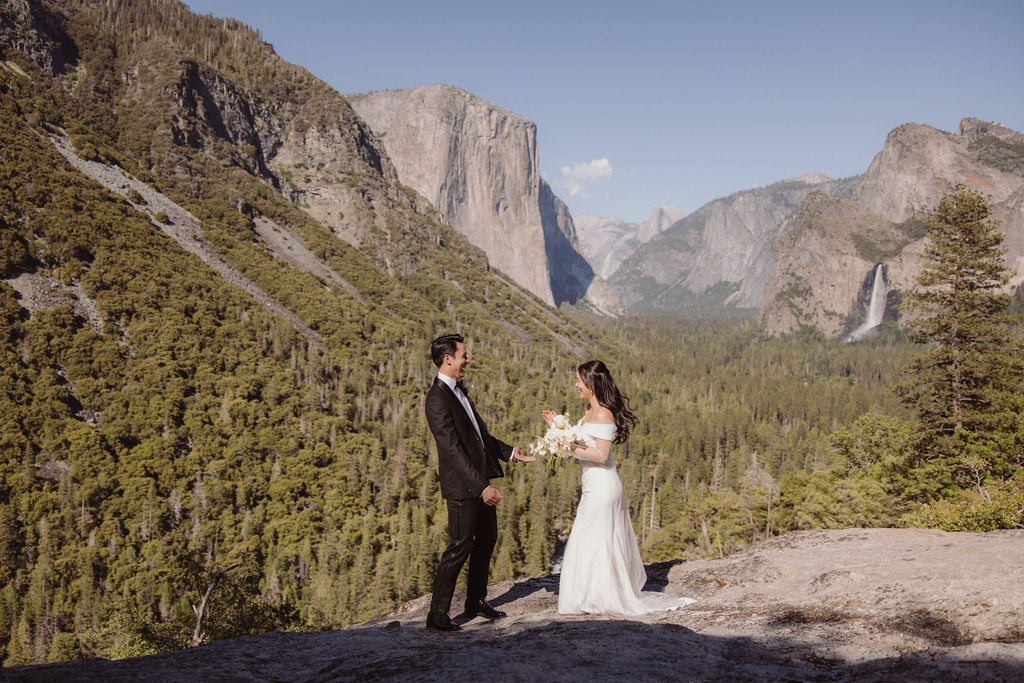 A bride in a white dress stands behind a groom in a black suit on a rocky overlook with mountains and trees in the background for their adventure elopement