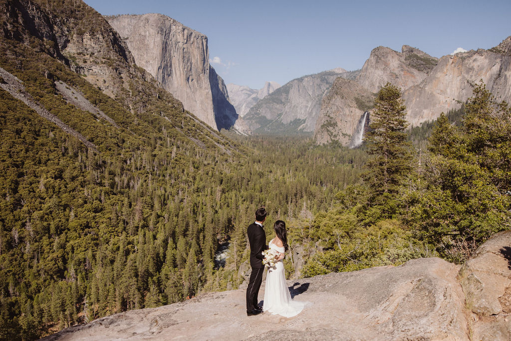 A bride in a white dress stands behind a groom in a black suit on a rocky overlook with mountains and trees in the background for their adventure elopement
