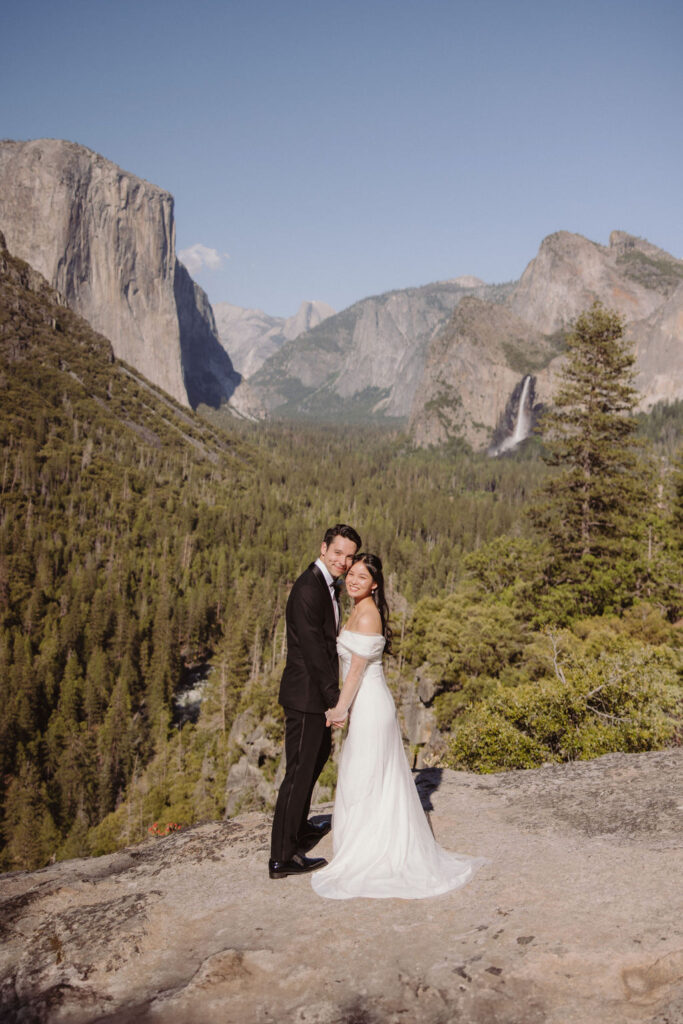 A bride in a white dress stands behind a groom in a black suit on a rocky overlook with mountains and trees in the background for their adventure elopement
