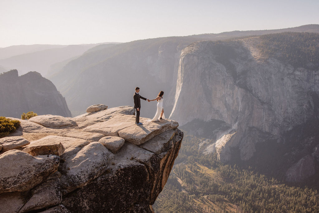A couple stands holding hands on the edge of a rocky cliff with a vast mountain landscape in the background.