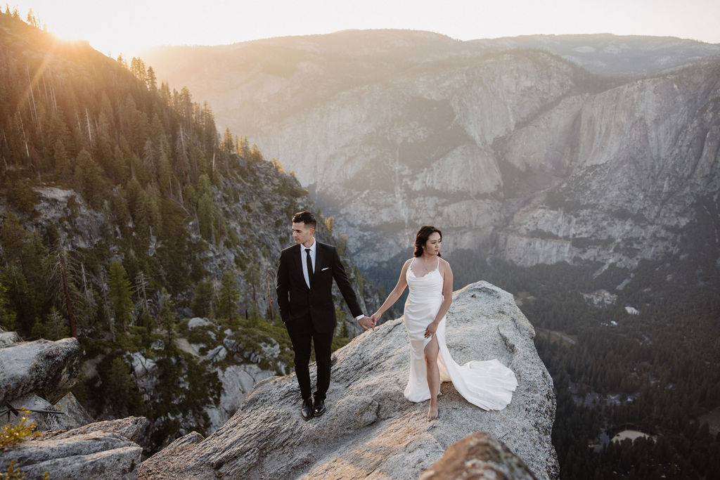 A bride and groom stand together on a rocky overlook with a mountain range and Half Dome in the background at sunset.