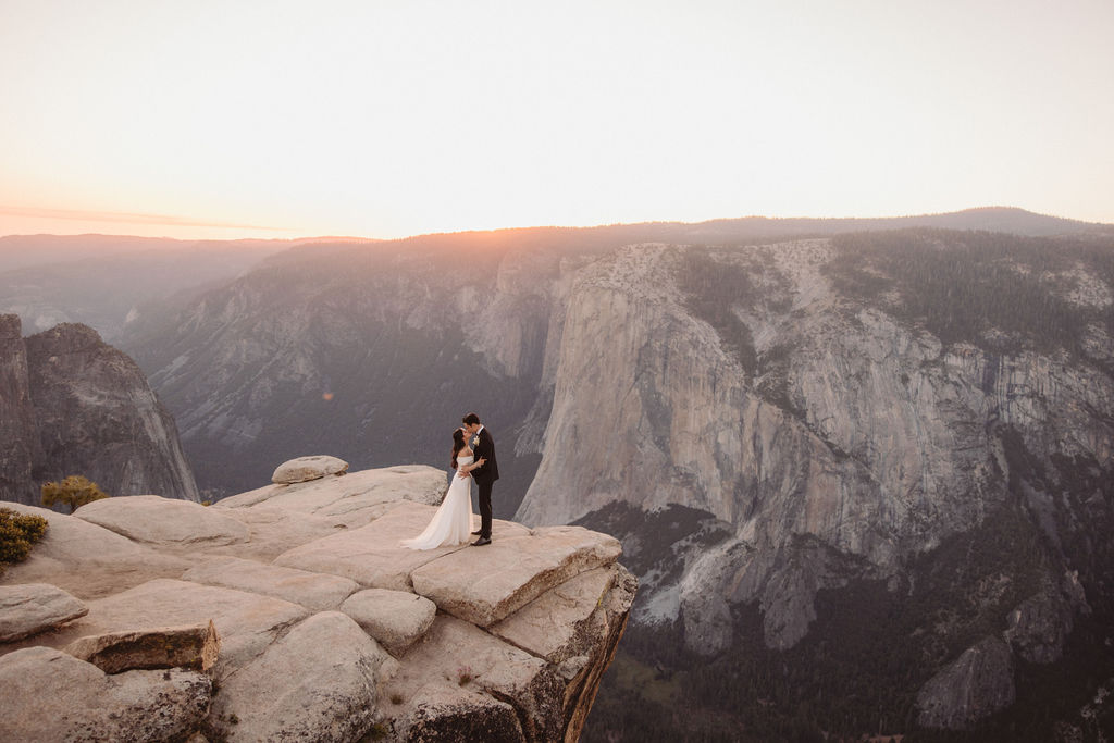 A bride and groom stand together on a rocky cliff edge at sunset, overlooking a vast mountainous landscape while they are eloping at a national park
