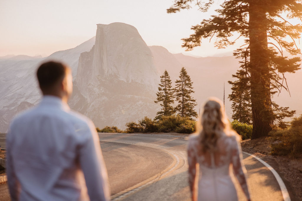A couple dressed in wedding attire walks hand in hand on a mountain road with Half Dome and pine trees in the background at sunset. | The Best Time for a Yosemite Elopement
