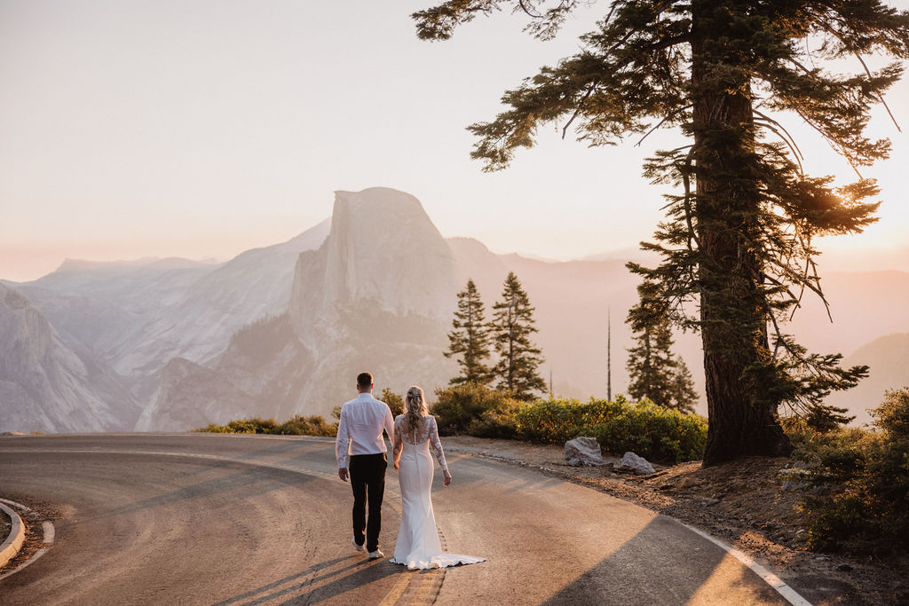 A couple dressed in wedding attire walks hand in hand on a mountain road with Half Dome and pine trees in the background at sunset. | The Best Time for a Yosemite Elopement