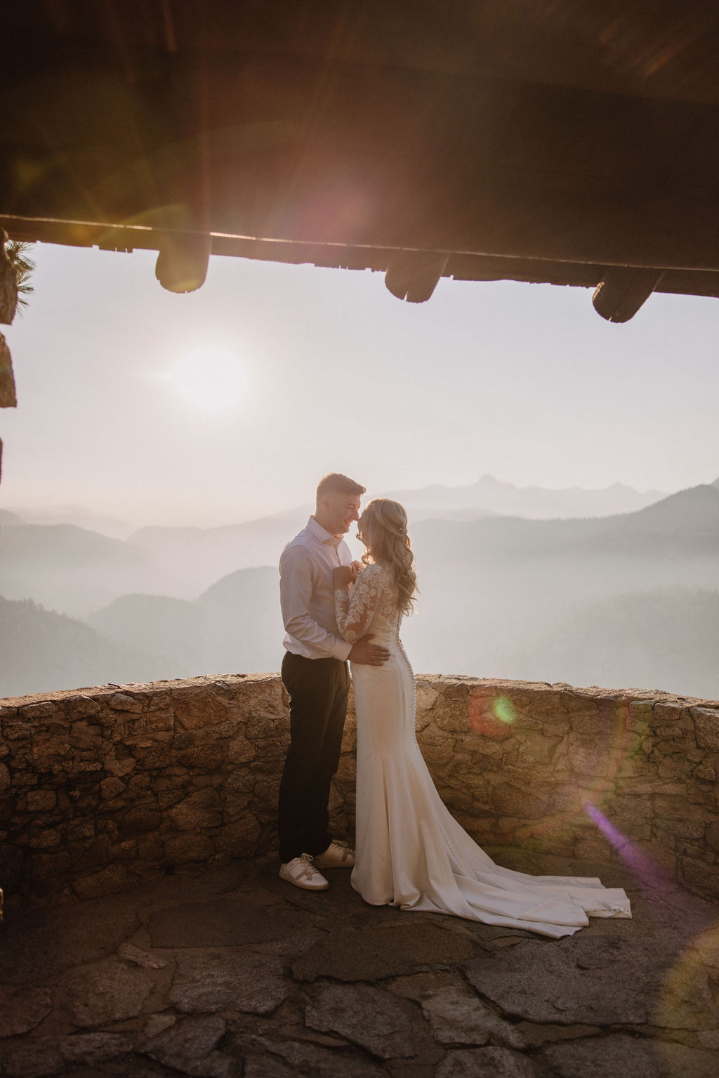 A couple dressed in wedding attire walks hand in hand on a mountain road with Half Dome and pine trees in the background at sunset. | The Best Time for a Yosemite Elopement