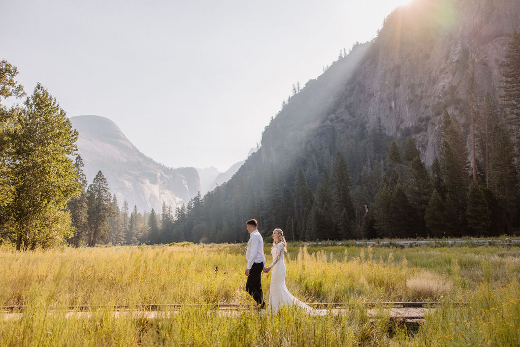 A couple in wedding attire stands holding hands on a wooden path in front of a grassy field and mountain, with sunlight streaming from behind.