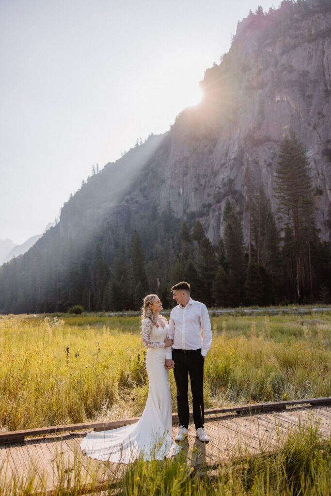 A couple in wedding attire stands holding hands on a wooden path in front of a grassy field and mountain, with sunlight streaming from behind.