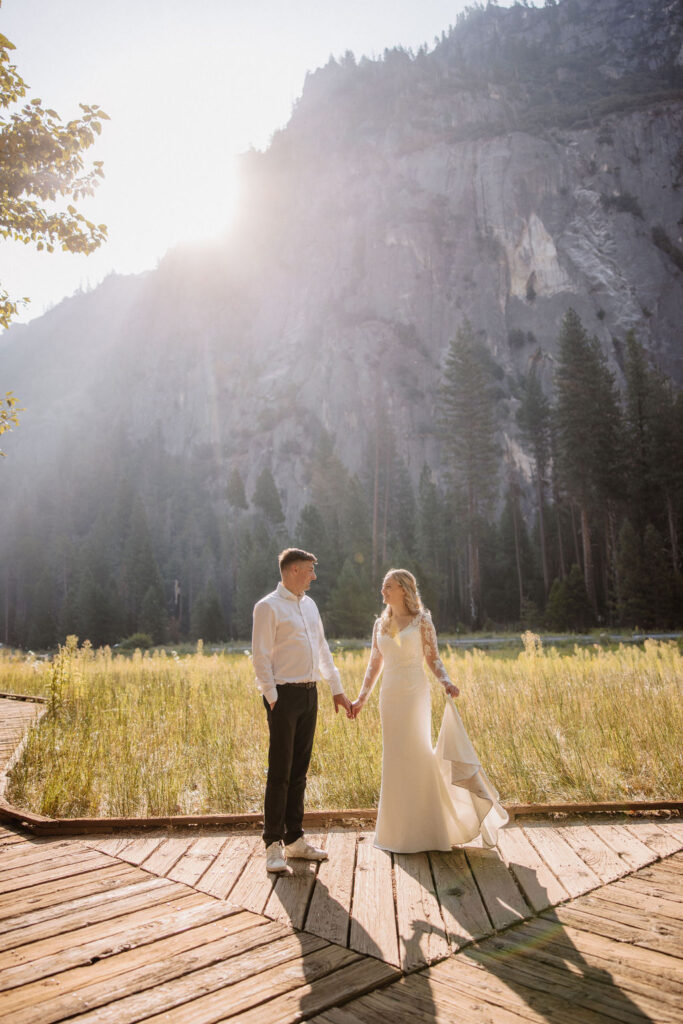 A couple in wedding attire stands holding hands on a wooden path in front of a grassy field and mountain, with sunlight streaming from behind.