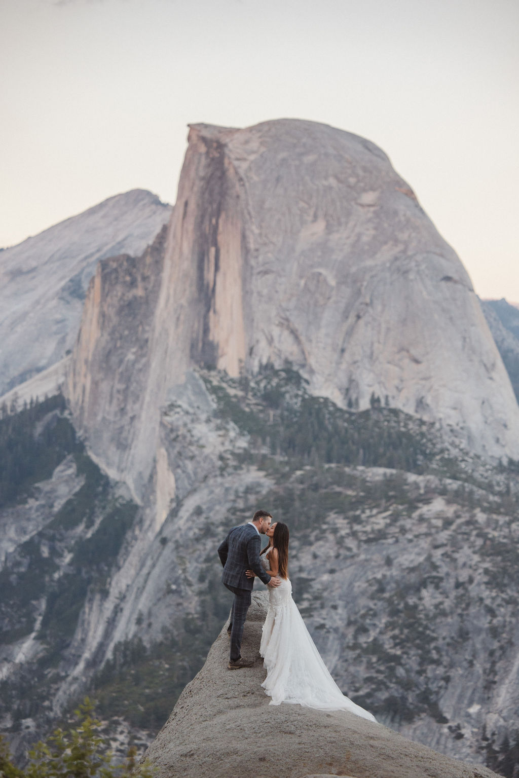 A bride and groom stand on a rocky ledge with Half Dome mountain in the background.