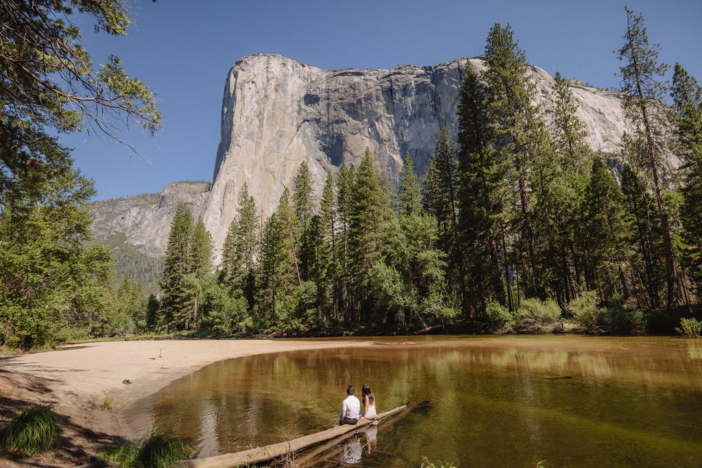 Two people sit on a log by a calm river, surrounded by pine trees, with a large rocky cliff rising in the background under a clear blue sky.