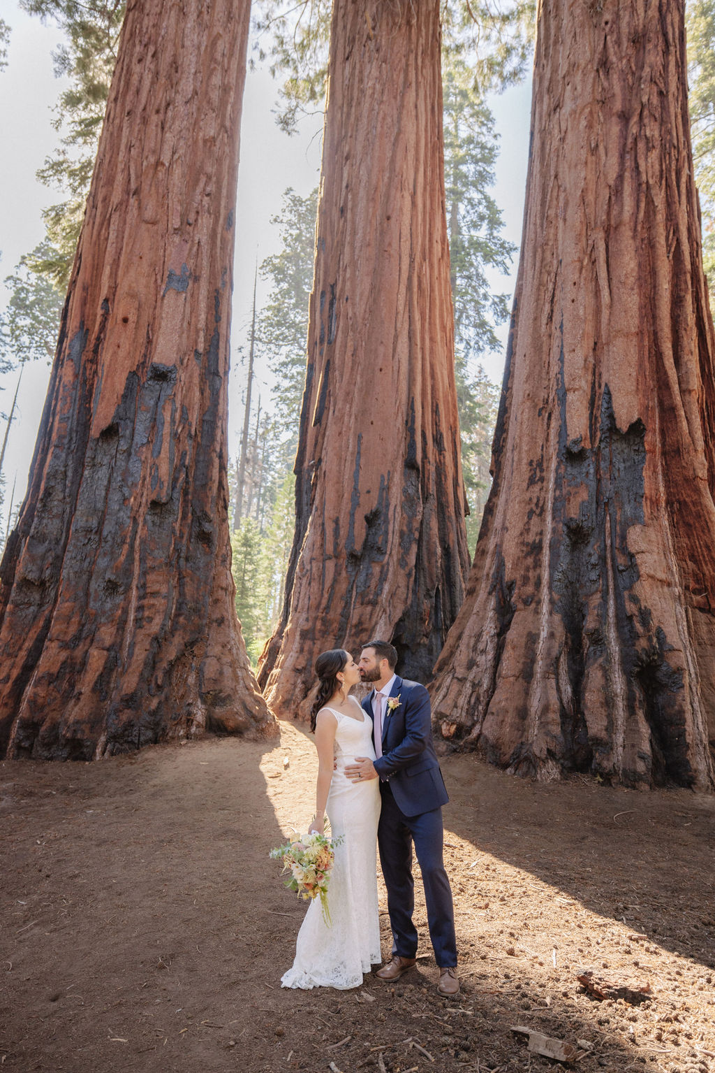 A couple stands inside the hollow base of a large sequoia tree in a forest, looking at each other as they elope in a national park