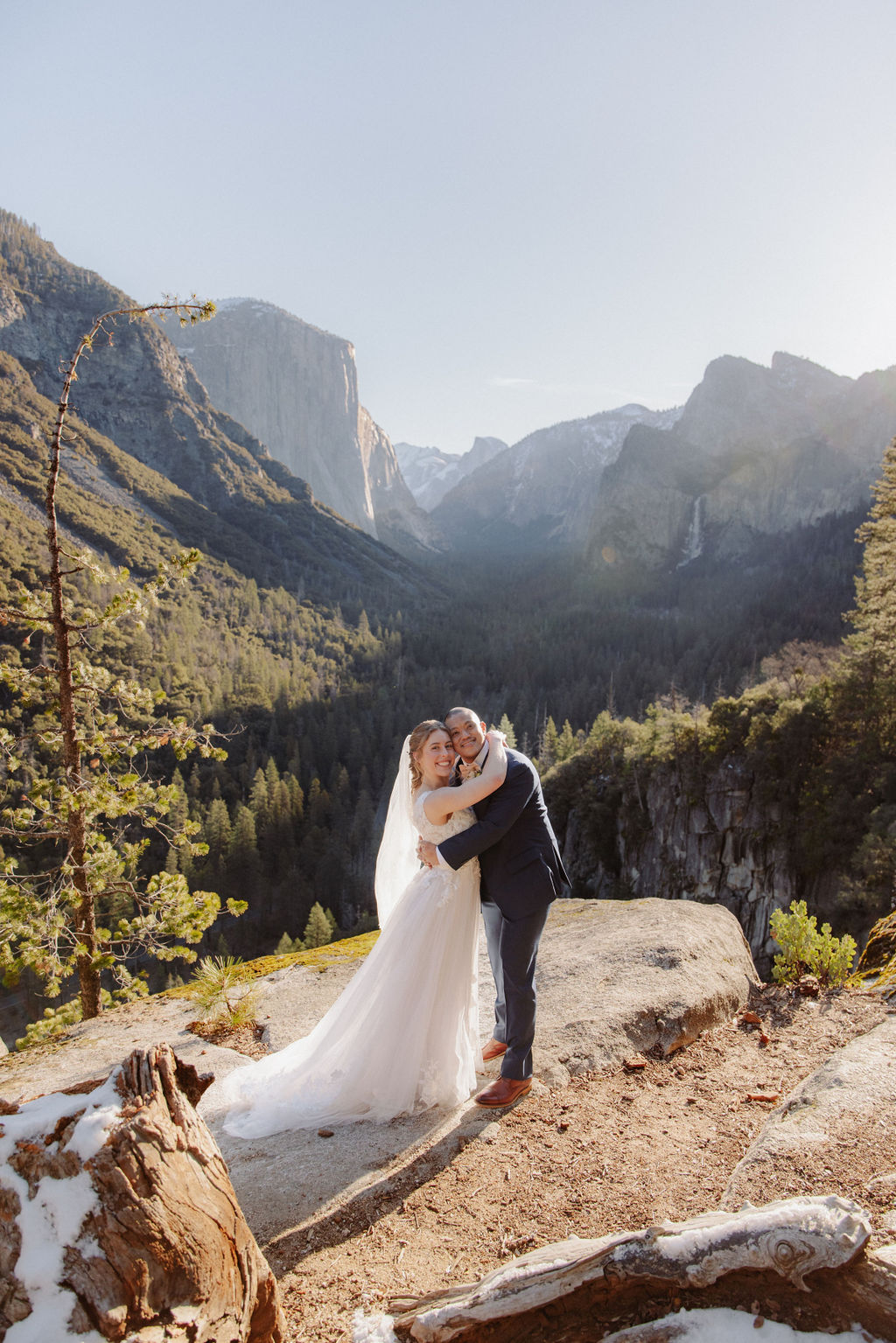 A bride and groom stand together on a rocky overlook with mountains and a forested valley in the background under clear skies.