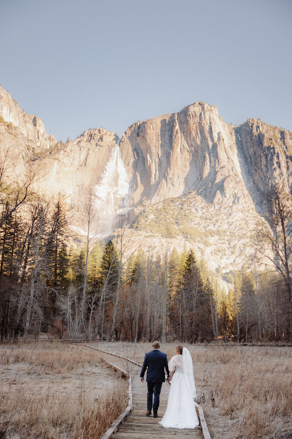 A bride and groom stand on a wooden pathway in a meadow with tall mountains and a waterfall in the background.