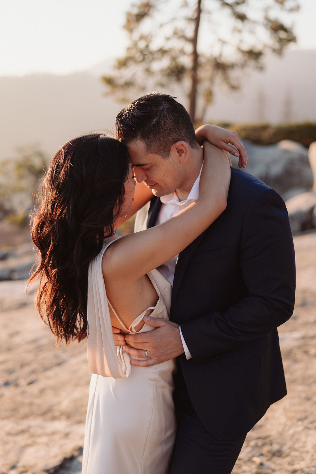 A couple in formal attire stands on rocky terrain at sunset, with mountains and trees visible in the background. | Elopement Planning Burnout Is Real & Here’s What to Do About It