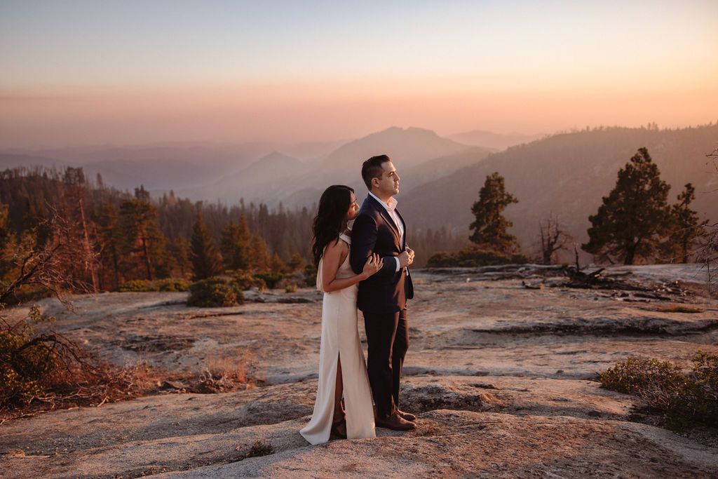 A couple in formal attire stands on rocky terrain at sunset, with mountains and trees visible in the background. | Elopement Planning Burnout Is Real & Here’s What to Do About It