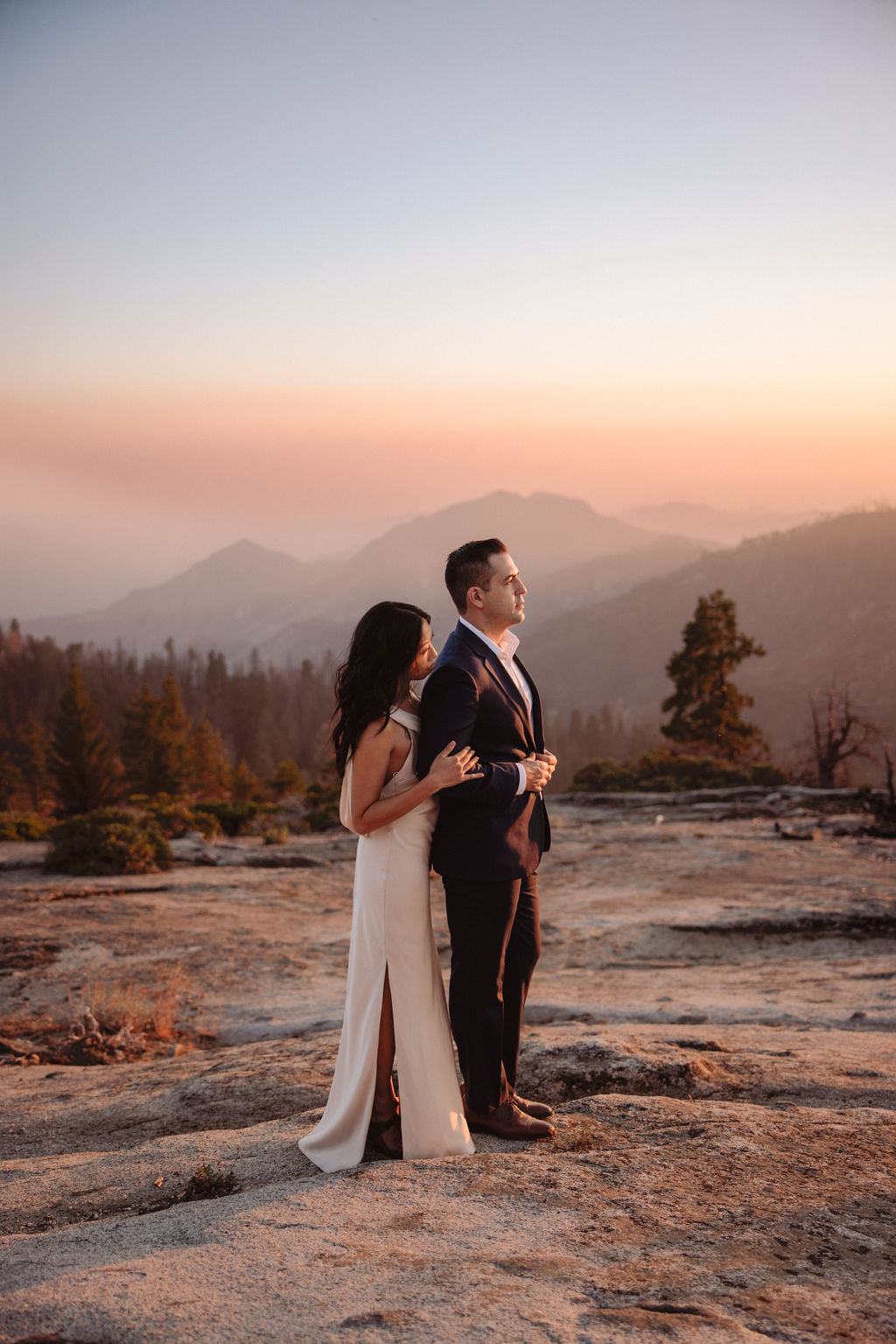 A couple in formal attire stands on rocky terrain at sunset, with mountains and trees visible in the background. | Elopement Planning Burnout Is Real & Here’s What to Do About It