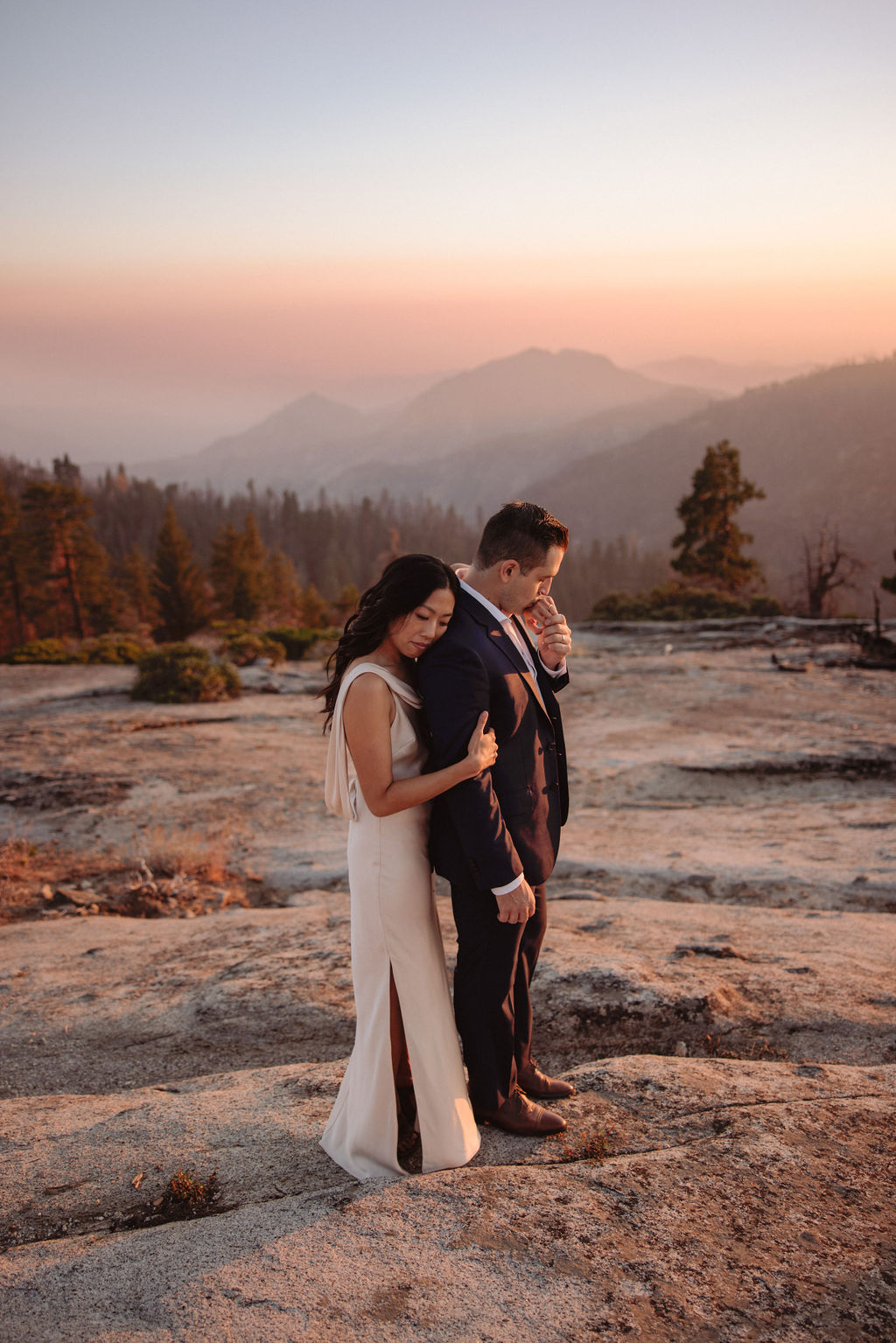 A couple in formal attire stands on rocky terrain at sunset, with mountains and trees visible in the background. | Elopement Planning Burnout Is Real & Here’s What to Do About It