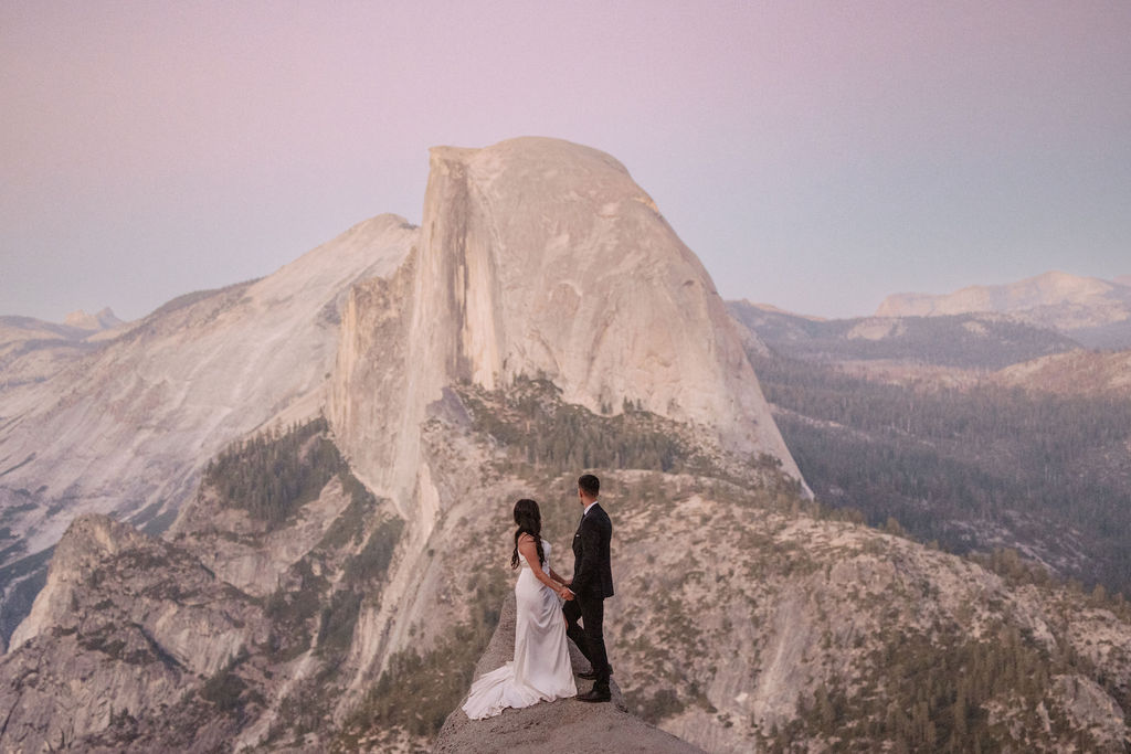 A bride and groom stand facing each other on a rocky ledge, exchanging vows, with a large mountain in the background for their adventure elopement