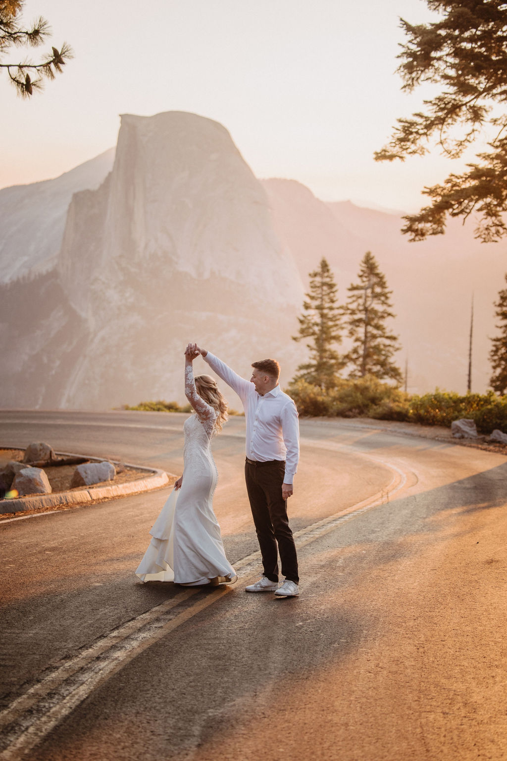 A couple dressed in wedding attire walks hand in hand on a mountain road with Half Dome and pine trees in the background at sunset. | The Best Time for a Yosemite Elopement
