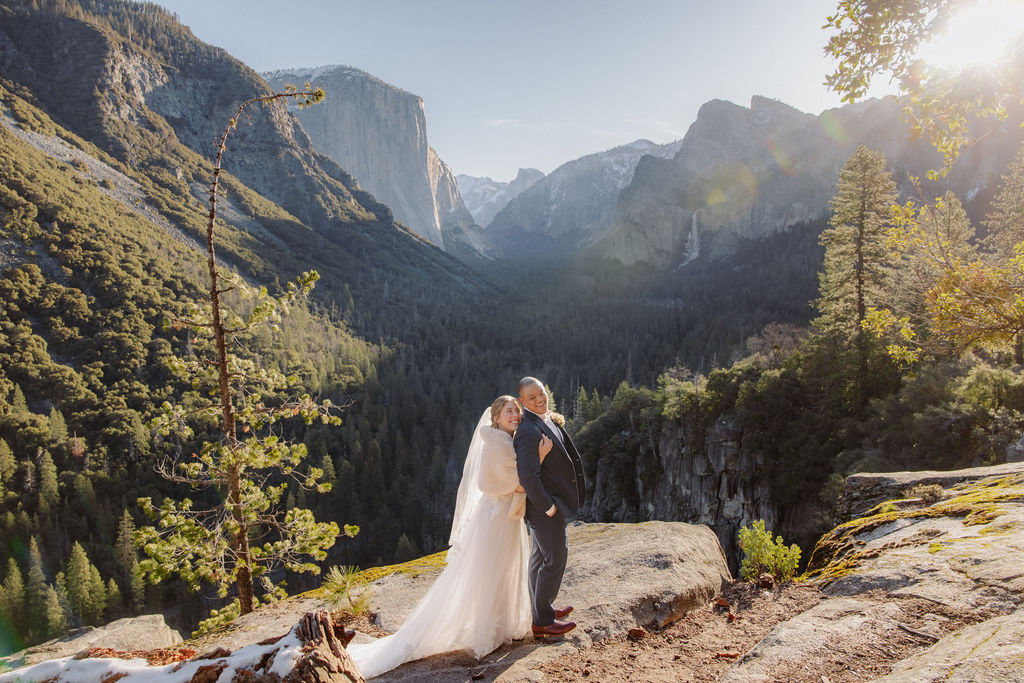 A bride and groom stand together on a rocky overlook with mountains and a forested valley in the background under clear skies.