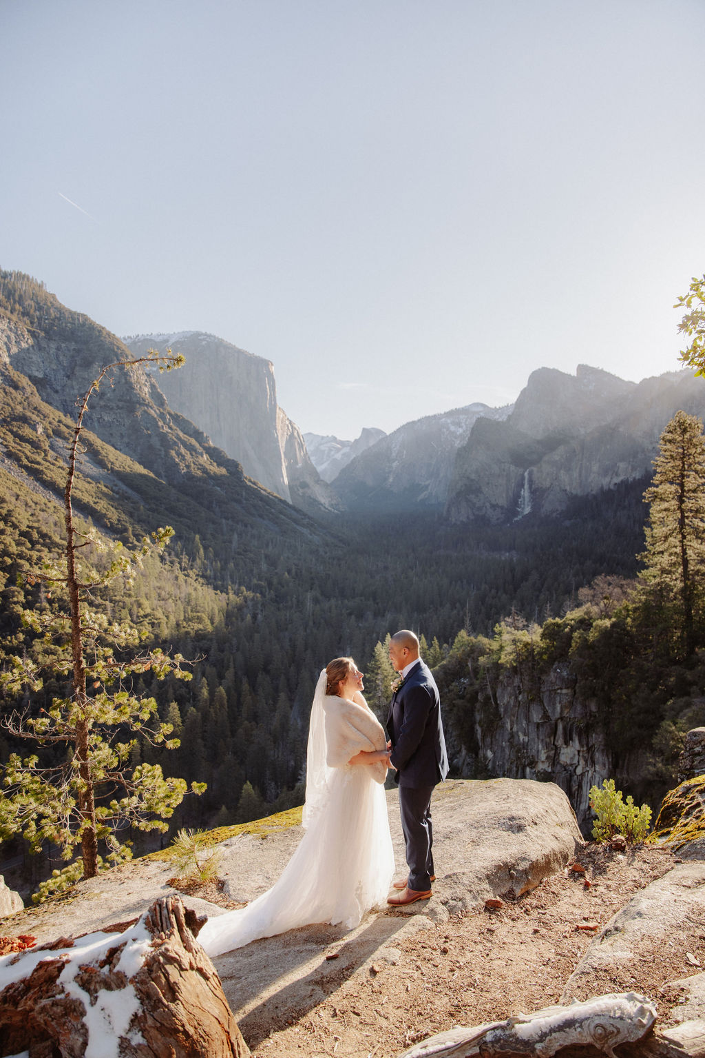 A bride and groom stand together on a rocky overlook with mountains and a forested valley in the background under clear skies.
