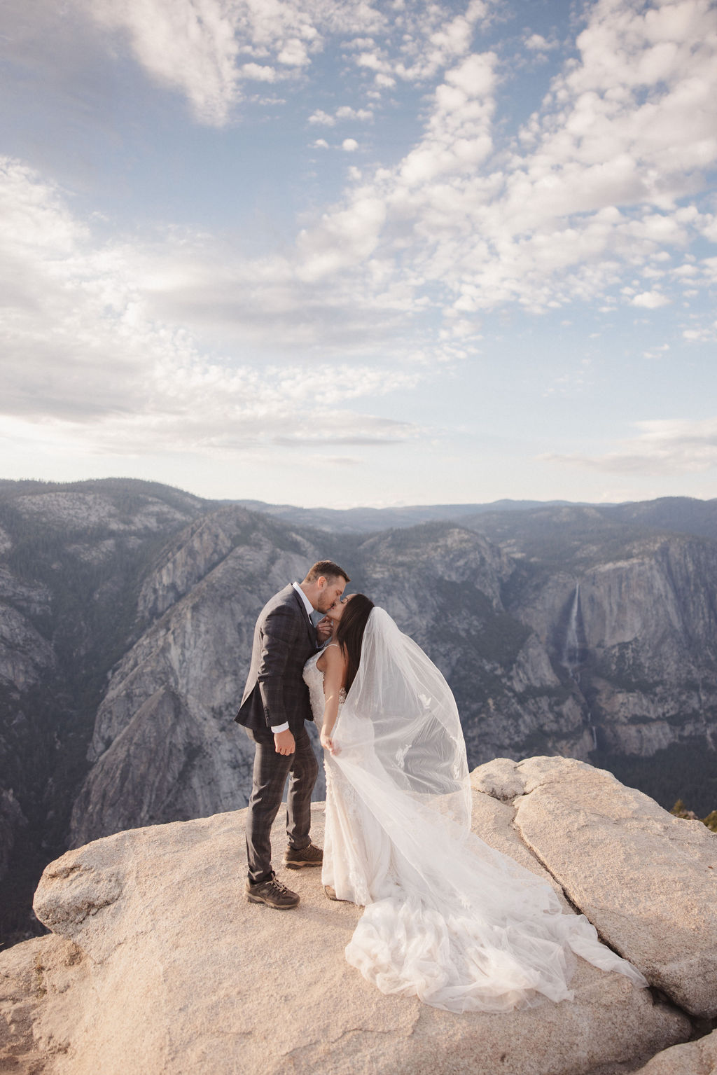 A bride and groom stand on a rocky ledge with Half Dome mountain in the background.