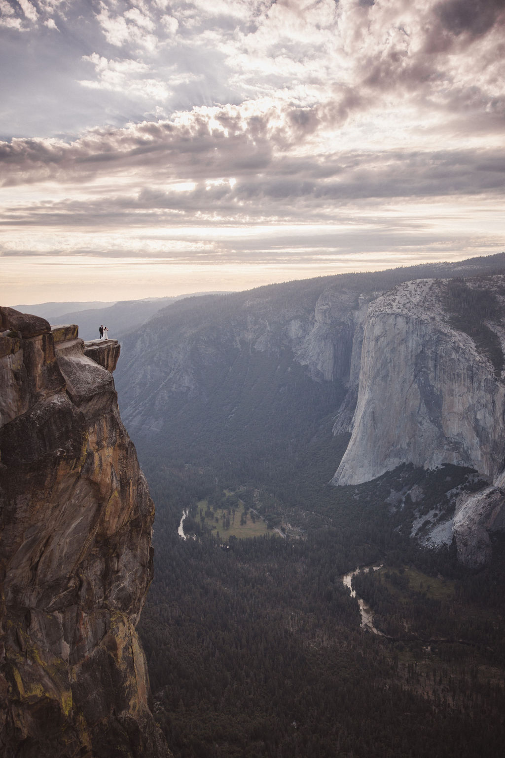 A bride and groom stand on a rocky ledge with Half Dome mountain in the background.