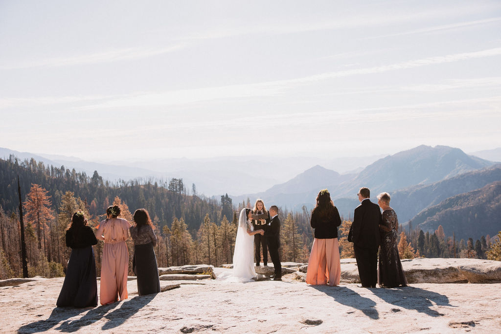 A small wedding ceremony takes place outdoors on a mountaintop, with guests standing and mountains visible in the background for their adventure elopement at sequoia national park
