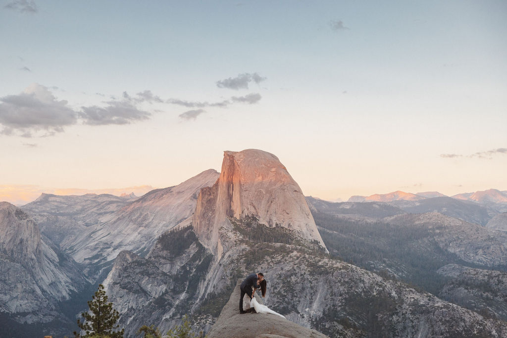 A bride and groom stand on a rocky ledge with Half Dome mountain in the background.