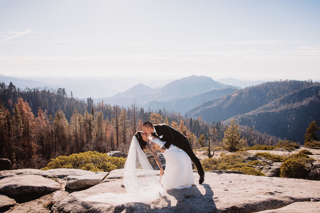 A small wedding ceremony takes place outdoors on a mountaintop, with guests standing and mountains visible in the background for their adventure elopement at sequoia national park