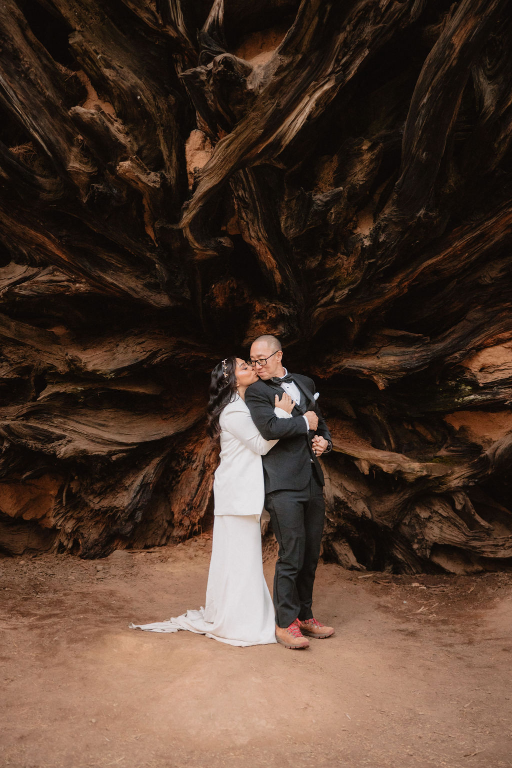 A couple in formal attire stands in front of the massive exposed roots of a fallen tree in a forest setting.