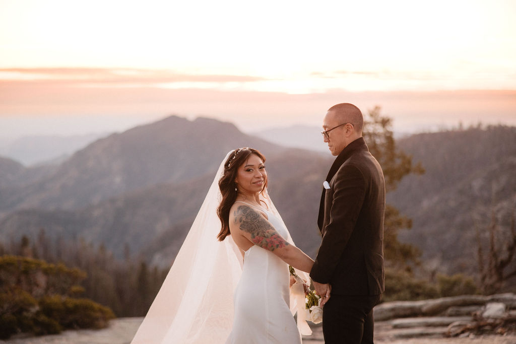A bride and groom stand together on a rocky landscape with mountains and trees in the background at sunset.