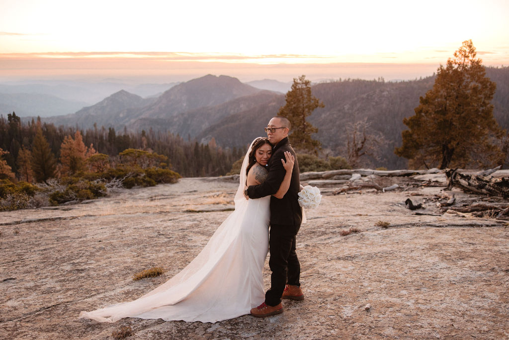 A bride and groom stand together on a rocky landscape with mountains and trees in the background at sunset.