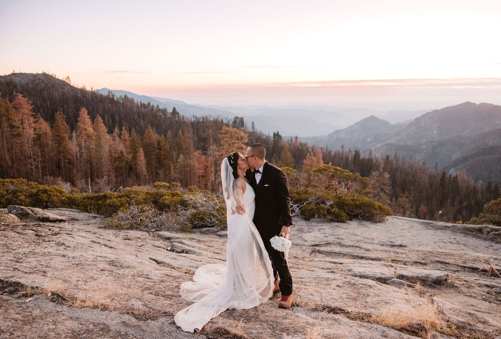 A bride and groom stand together on a rocky landscape with mountains and trees in the background at sunset.